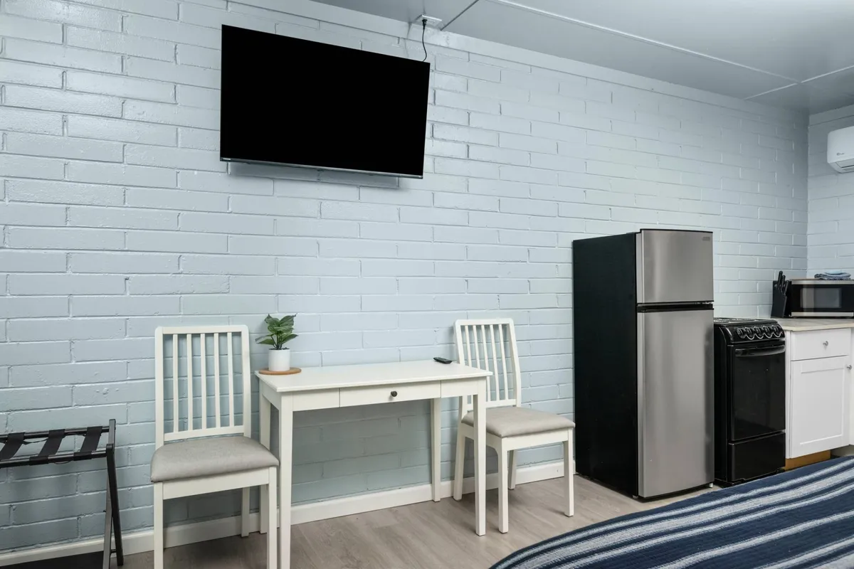 A functional and inviting living space featuring a TV mounted on the wall, a small table with two chairs, a refrigerator, a stove, and a decorative plant. The decor is modern and neutral, with furnishings that look both comfortable and stylish.