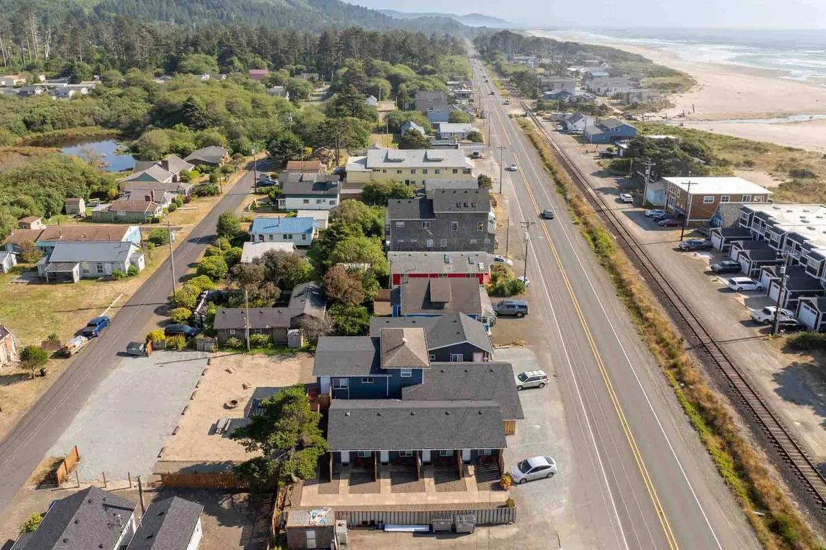 A broad exterior scene showing the beach, a community road, houses nestled among lush vegetation, and people engaged in walking, biking, and gathering. The relaxed mood captures the essence of a beach town where travel by foot or bike is common and enjoyable.