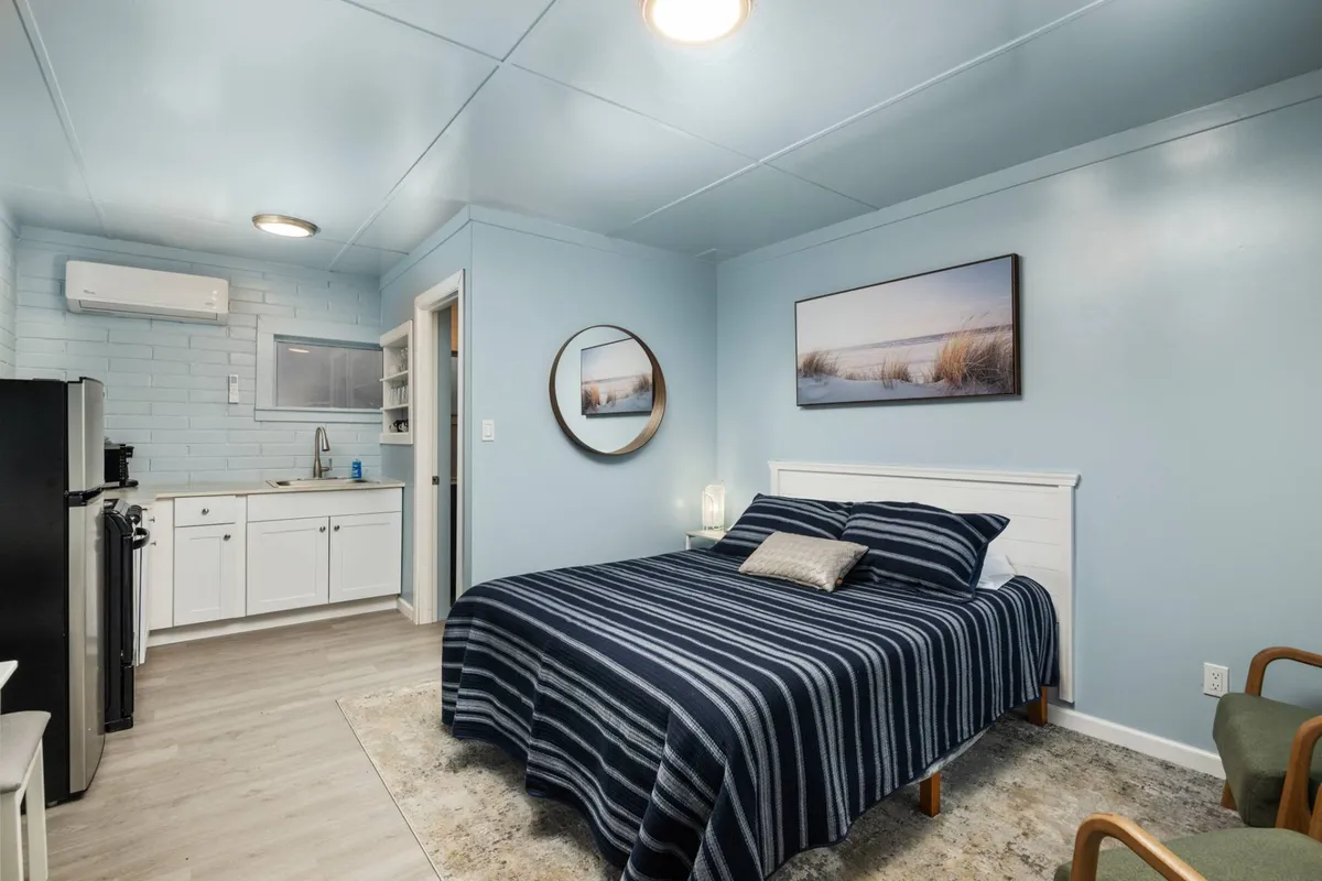 A tranquil bedroom with a bed featuring striped bedding, framed artwork above the headboard, a round mirror, and a compact kitchenette area in the background. Soft natural light filters in, enhancing the calm and comfortable atmosphere.