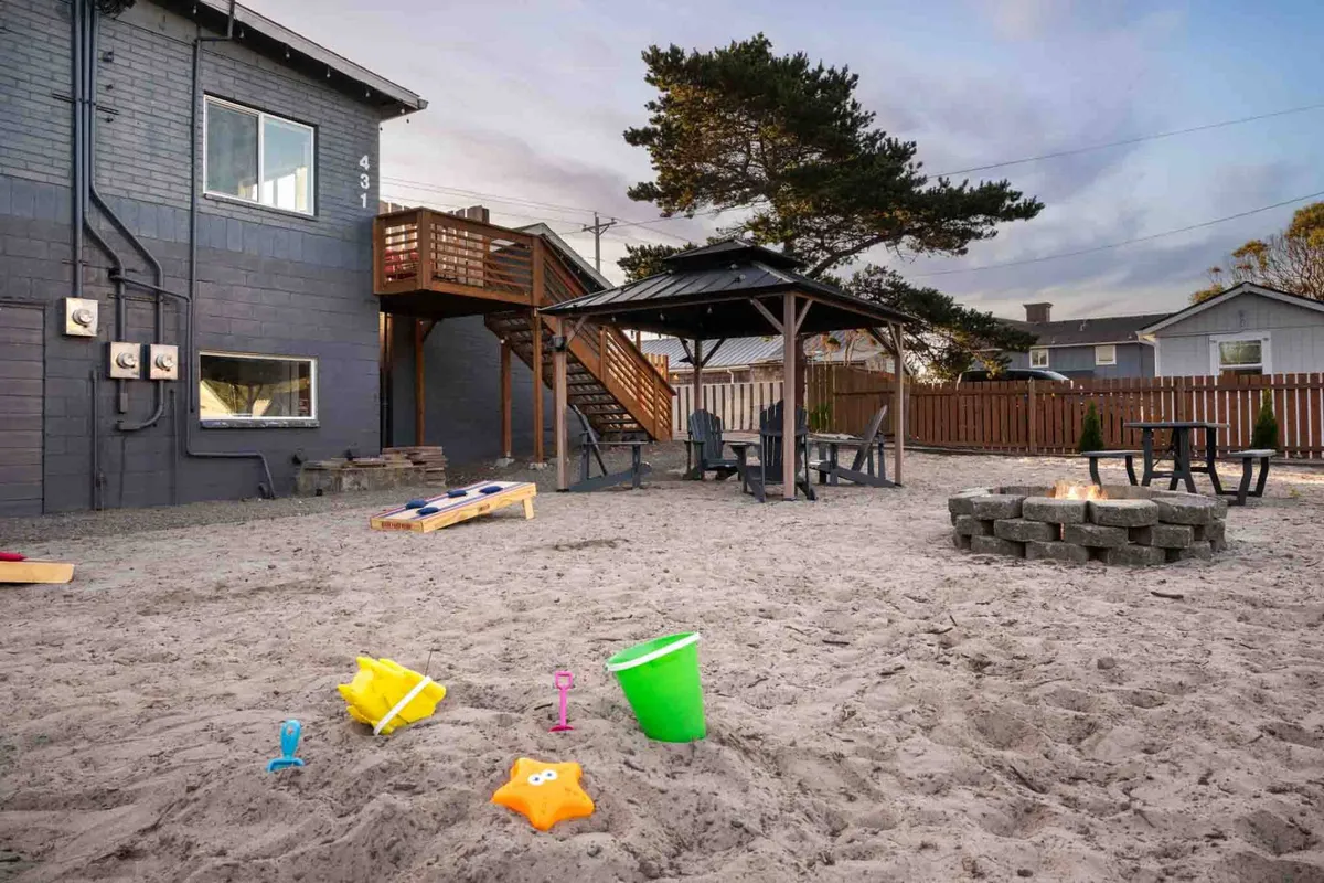 This image features a shared gazebo with outdoor seating, a fire pit, and playful beach toys scattered in the sand. The scene is lively, showing both adults and children enjoying communal activities, including family gatherings, evening bonfires, and socializing with friends under the gazebo.