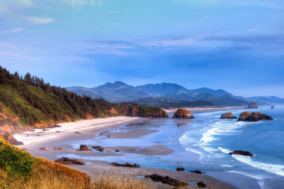 A scenic view showing the sandy expanse of Rockaway Beach, with rocky outcrops and gentle waves crashing ashore. In the distance, misty mountains frame the horizon, creating a serene and atmospheric setting. The beach looks inviting for walks, exploration, and relaxation.