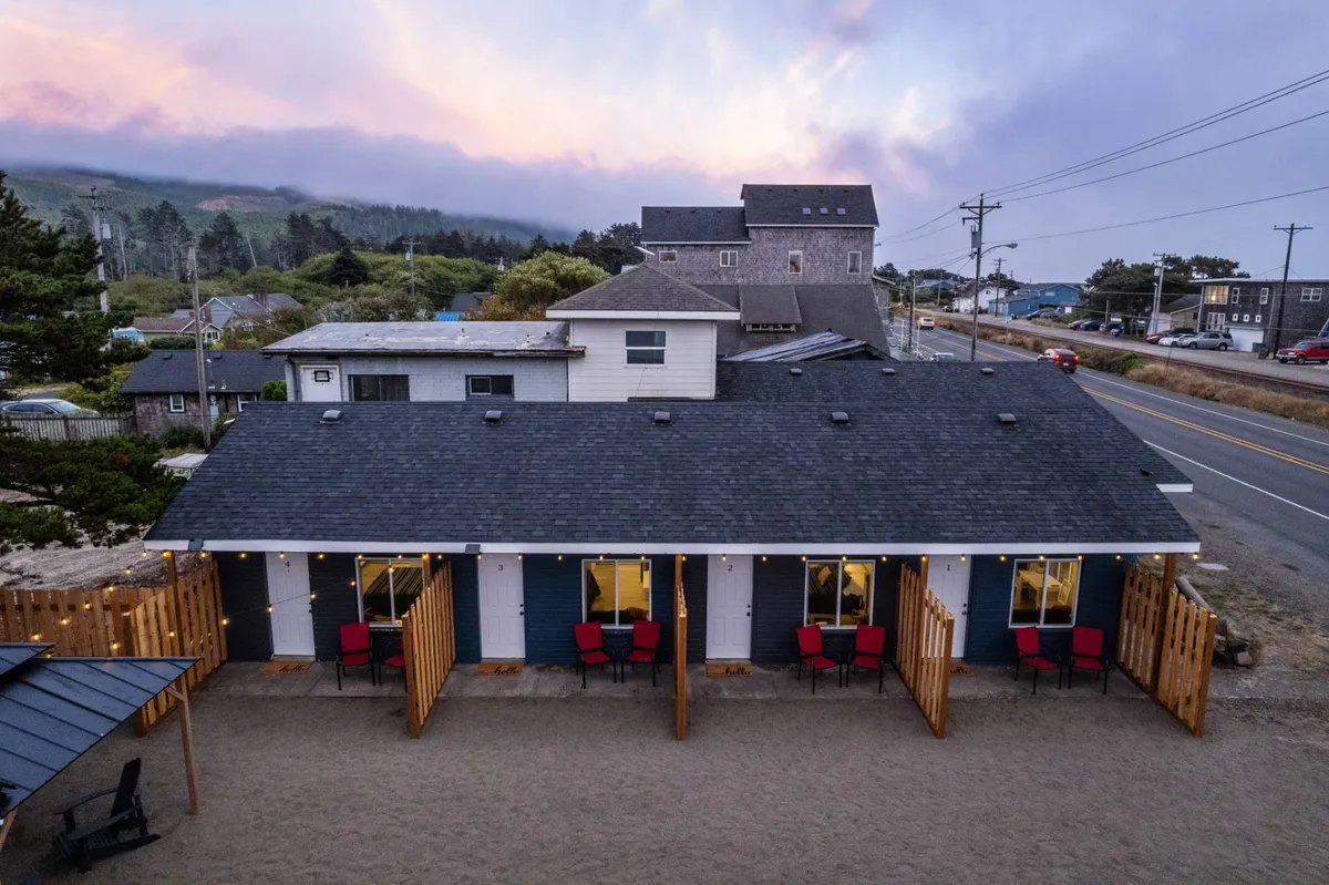 A wooden deck with bright red chairs set against the cottage exterior, windows glowing softly from interior lighting. The deck overlooks a sandy area, suggesting a seamless transition from the private outdoor space to the surrounding natural environment.