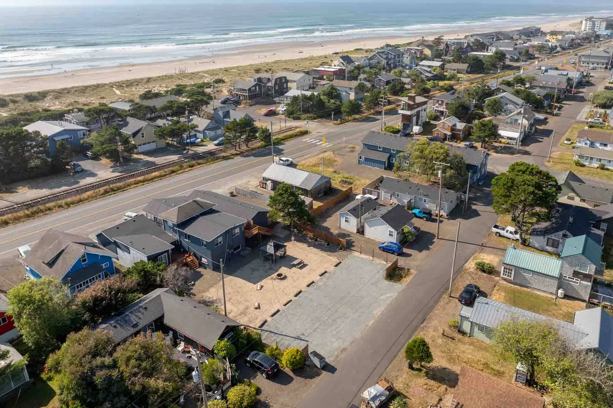 An inviting exterior shot with the sandy beach, a few houses set back from the shore, a bordering road, and lush greenery. The scene conveys a relaxed, walkable environment ideal for sunbathing, building sandcastles, or exploring tide pools.