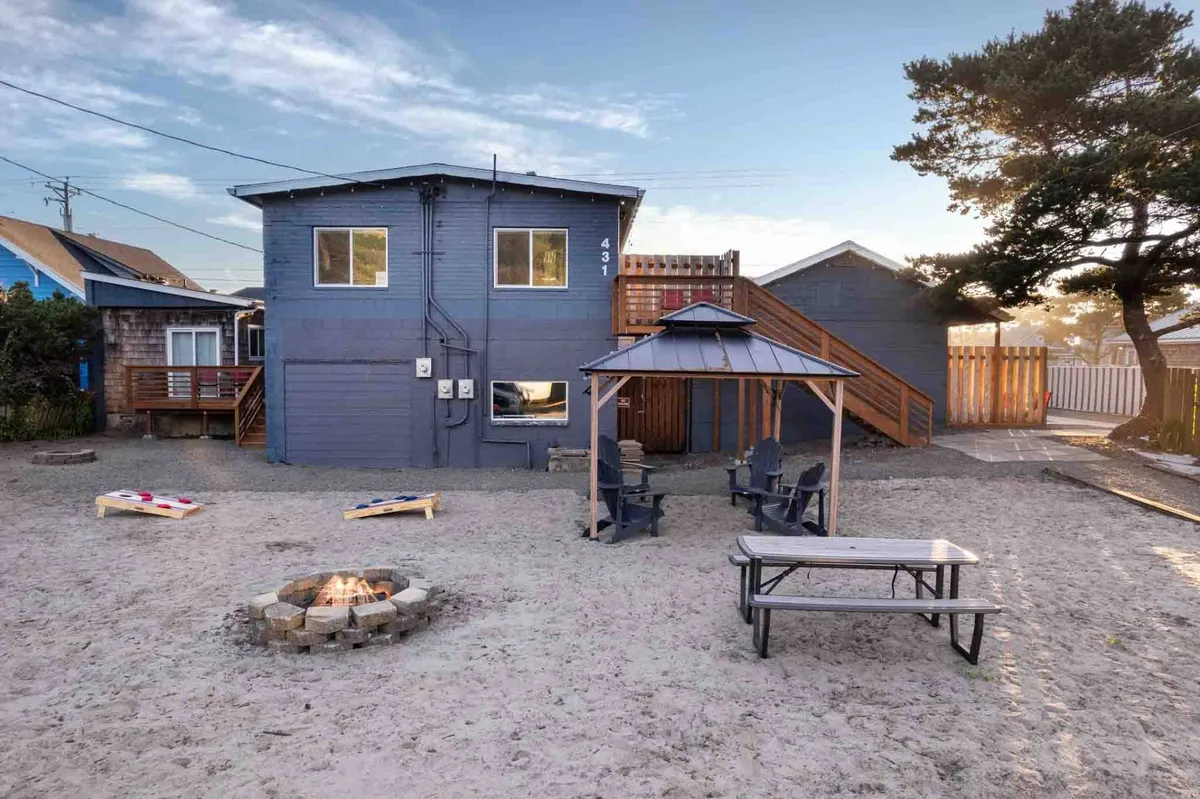 The photo captures an inviting outdoor setting with a gazebo, fire pit, several outdoor seating options, and a cornhole game set up nearby. People are seen relaxing and socializing, enjoying both games and fireside chats in a cozy, communal setting.