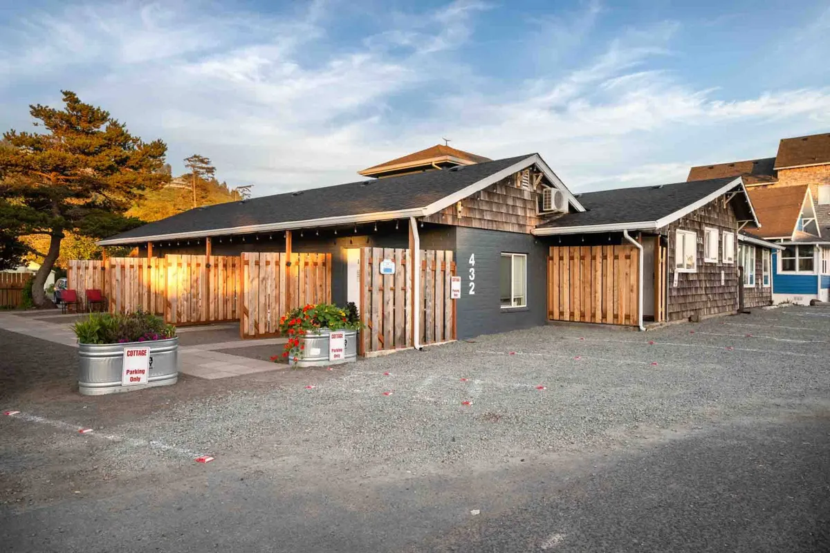 This exterior photo features the inviting wooden siding of a cottage surrounded by flower beds, a visible parking area, and signage that hints at a welcoming community atmosphere. The scene suggests easy access to the outdoors and nearby attractions, with subtle cues of people relaxing or gathering just out of frame.