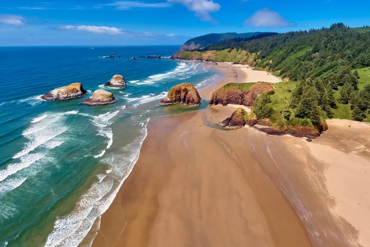 A sweeping view of the nearby beach, with waves crashing onto a wide stretch of sandy shore, rock formations creating visual interest, and lush green hills rising in the background. The scene is tranquil and inviting, perfect for outdoor exploration and leisurely walks by the sea.