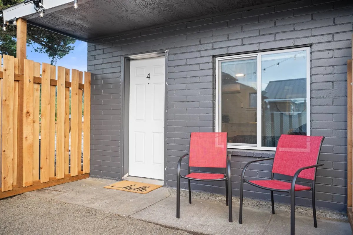 A private cottage exterior showing a door marked with the number 4, flanked by two inviting red chairs on a small patio, bordered by a wooden fence and a welcome mat at the entrance. The patio appears intimate and cozy, ideal for sitting outdoors in privacy.