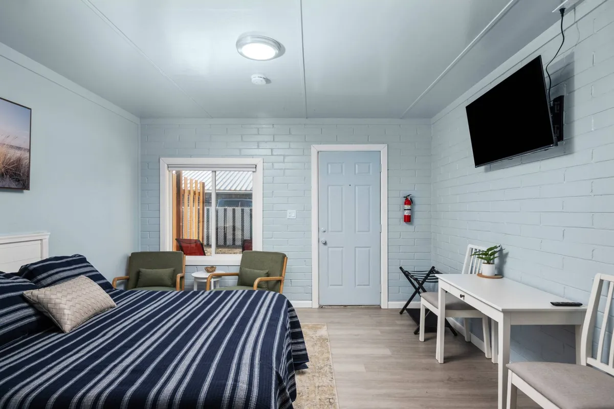 A welcoming bedroom scene with a bed dressed in striped bedding, a window offering outdoor views, a wall-mounted TV, and a small desk with a chair. The space is bathed in natural light, inviting relaxation or a peaceful morning routine.